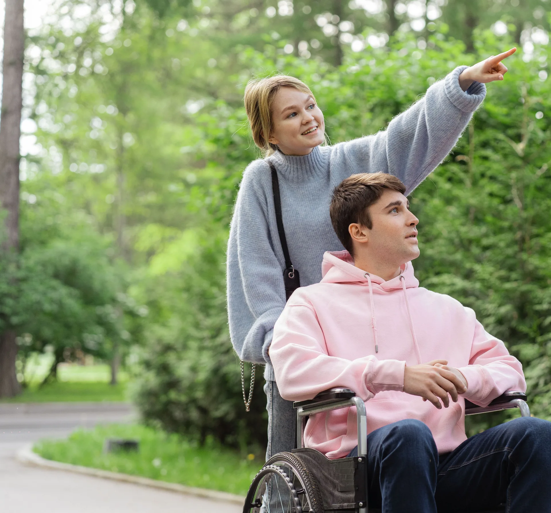 Een begeleider en een jongeman in een rolstoel lachen samen buiten in een groen park; zij wijst iets aan