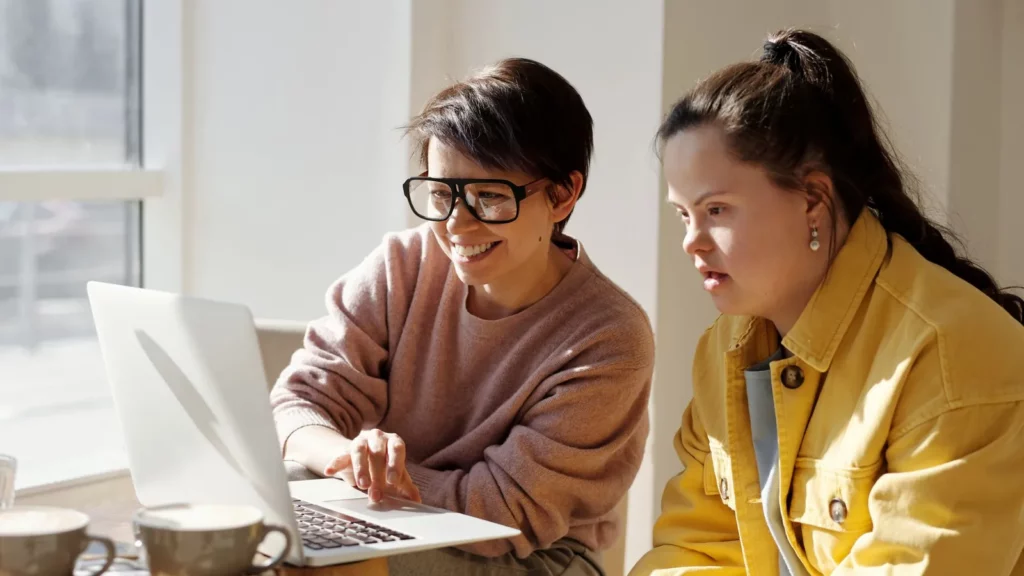 Twee vrouwen kijken samen op een laptop aan tafel bij het raam; de vrouw rechts heeft het syndroom van Down