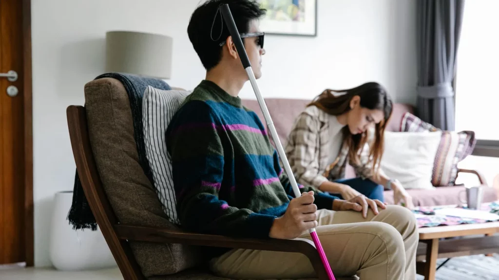 Een man met een taststok zit in een fauteuil; een vrouw zit naast hem en leest mee