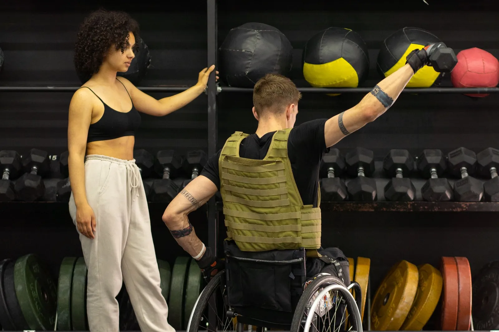 Een vrouw begeleidt een man in een rolstoel bij een oefening in een sportzaal